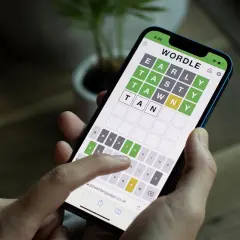 Dłoń trzyma smartfon z grą typu wordle. Na ekranie widać planszę z literami i częściowo odgadnięte słowo.
