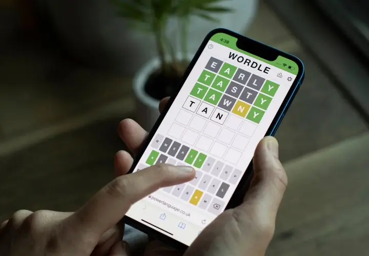 Dłoń trzyma smartfon z grą typu wordle. Na ekranie widać planszę z literami i częściowo odgadnięte słowo.