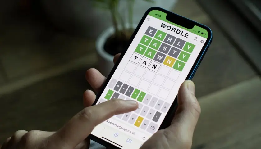 Dłoń trzyma smartfon z grą typu wordle. Na ekranie widać planszę z literami i częściowo odgadnięte słowo.