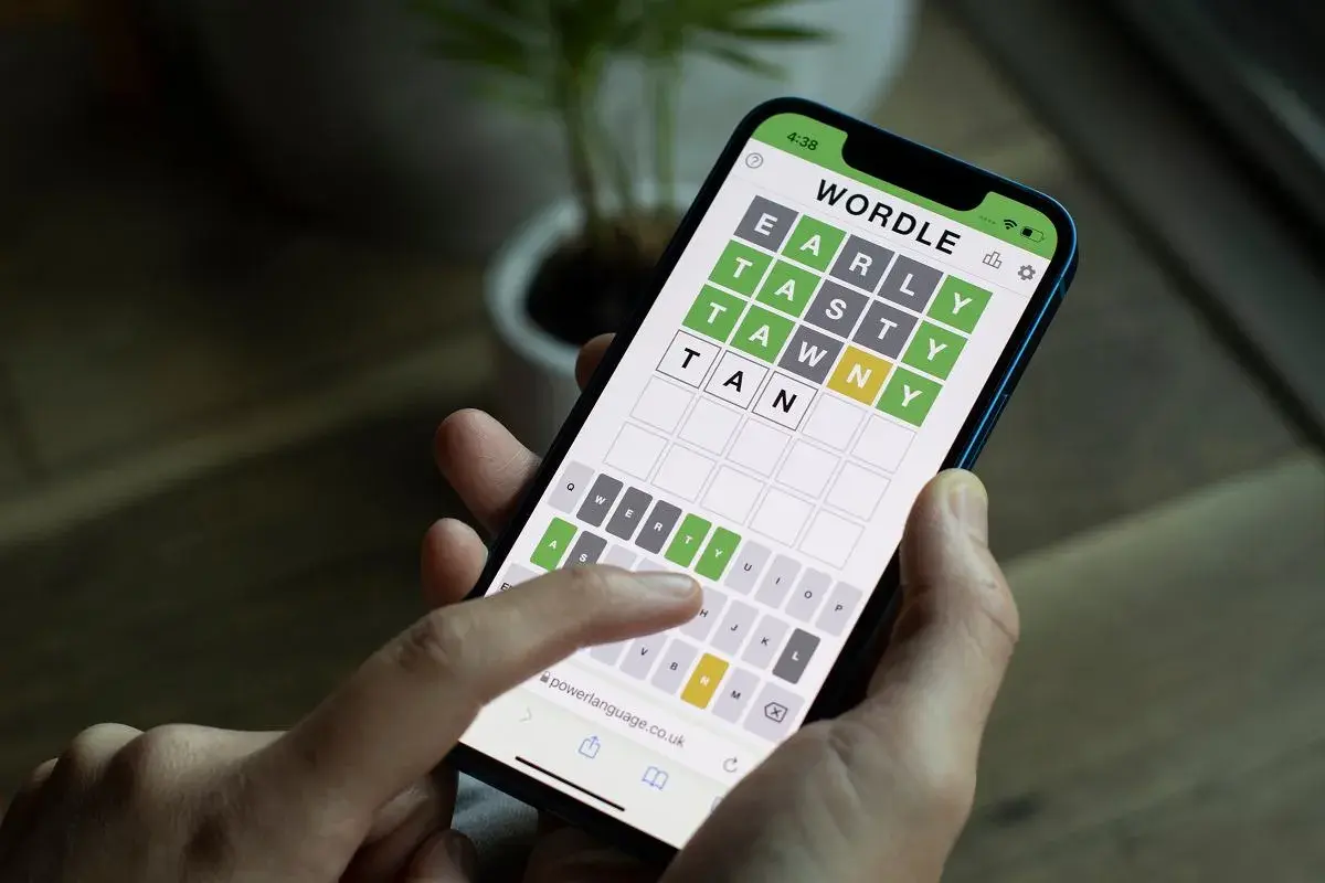 Dłoń trzyma smartfon z grą typu wordle. Na ekranie widać planszę z literami i częściowo odgadnięte słowo.