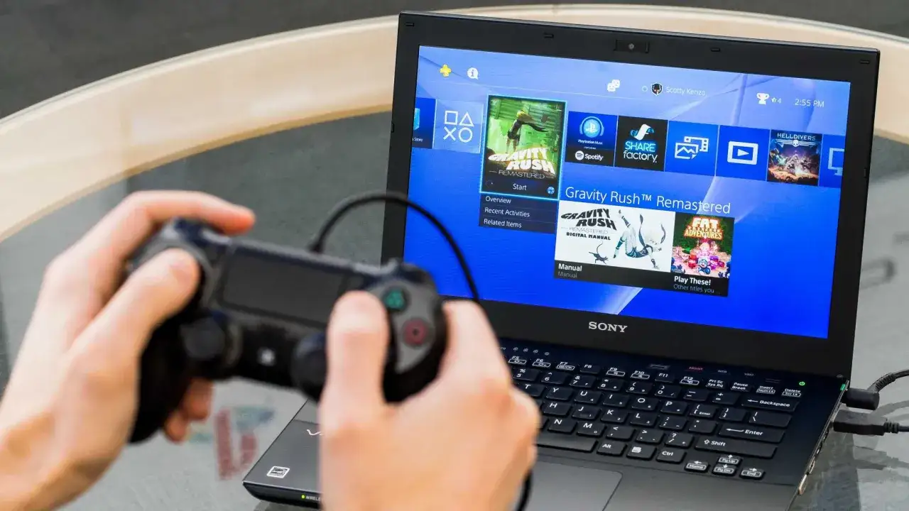 Gracz steruje grą na laptopie przez PS Remote Play PC. Na ekranie widać menu PlayStation z grą Gravity Rush Remastered.
